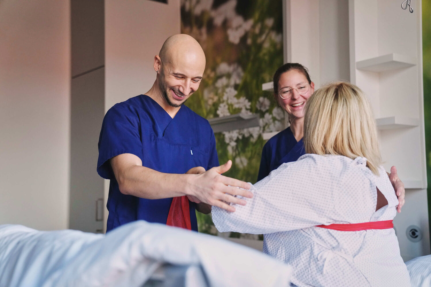 Das Bild zeigt einen pflegerischen Mitarbeiter und eine Mitarbeiterin der Pflege im Gespräch mit einer im Bett sitzenden Patientin.