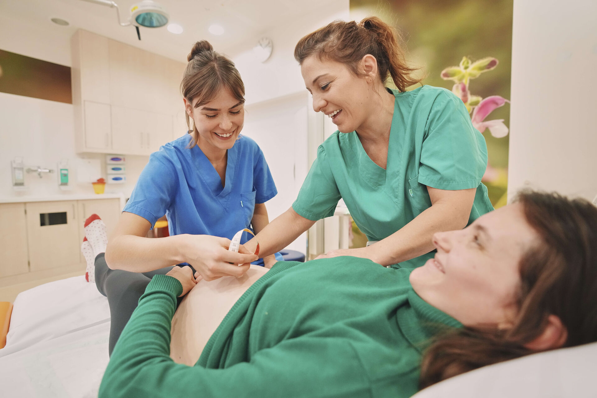 Das Bild zeigt zwei lächelnde Mitarbeiterinnen der Sana Kliniken mit einer liegenden Patientin.